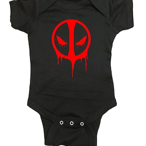 May include: Black baby onesie with a red Deadpool logo. The logo is a circle with a red mask and dripping red paint.