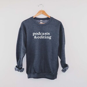 K&ouml;nnte beinhalten: Ein dunkelgrauer Pullover mit dem Aufdruck "podcasts & editing" in Wei&szlig; auf der Vorderseite.