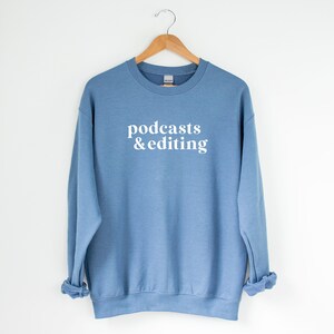 K&ouml;nnte beinhalten: Ein hellblauer Pullover mit dem Aufdruck "podcasts & editing" in Wei&szlig; auf der Vorderseite.