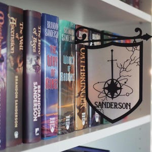 May include: A bookshelf with a collection of fantasy novels by Brandon Sanderson. The books are arranged in order of publication, with the most recent book on the right. The books are all hardcovers and have a similar design. The bookshelf is made of wood and has a white finish. A black metal sign with a compass and sword design hangs on the shelf. The sign reads "Sanderson".