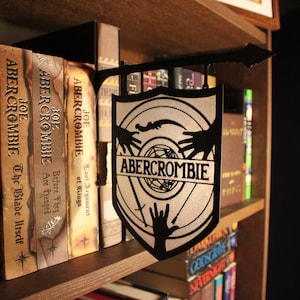Puede incluir: Un letrero de metal negro con fondo plateado y el texto "ABERCROMBIE" en un círculo blanco. El letrero está colgado en un estante de madera con libros detrás.