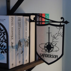 May include: A bookshelf with a collection of fantasy novels by Brandon Sanderson. The books are titled "The Final Empire", "The Well of Ascension", and "Hero of Ages". A metal sign with the text "Sanderson" and a compass design hangs from the shelf.