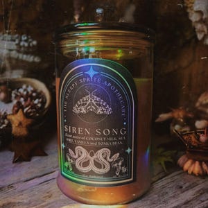 May include: A glass jar candle with a black label featuring a moth illustration and the text "SIREN SONG". The label also lists scent notes of coconut milk, sea salt, vanilla, and tonka bean. The candle has a lid and a warm, amber glow.