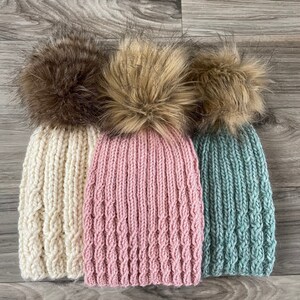 May include: Three knitted hats with faux fur pom poms. The hats are in cream, pink, and teal.