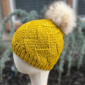 May include: A mustard yellow knit beanie with a textured pattern and a fluffy, cream-colored pom-pom on top. The hat is displayed on a mannequin head, showcasing its shape and design. The beanie is a winter accessory.