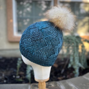 May include: A teal blue knitted beanie with a white pom pom. The beanie has a textured knit pattern.