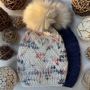 May include: A cozy, hand-knitted winter hat with a large, fluffy, light brown pom-pom. The hat features a textured, off-white pattern with flecks of gray and orange. A second, solid navy blue hat is visible next to it.