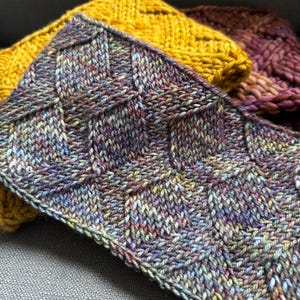 Op de afbeelding: Close-up van een gebreide stof met een textuurpatroon in tinten paars, blauw en groen.