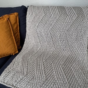 Puede incluir: Una manta gris tejida con un patrón de espiga está sobre un sofá azul oscuro. Dos cojines naranjas son visibles en el lado izquierdo. La textura de la manta es claramente visible.