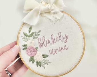 May include: A white embroidery hoop with a pink floral design and the name "Blakeily Anne" embroidered in pink thread.