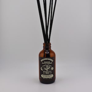 May include: An amber glass reed diffuser bottle with black reeds. The label reads Project Happiness Reed Diffuser, Made With Clean Essential Oils, Peppermint & Eucalyptus, 3.5 fl oz / 103 ml. The bottle has a narrow neck and a cylindrical body.