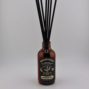 May include: An amber glass reed diffuser bottle with black reeds. The label reads Project Happiness Reed Diffuser, Made With Clean Essential Oils, Mango & Coconut Milk, 3.5 fl oz / 103 ml. The bottle has a narrow neck and a black cap.