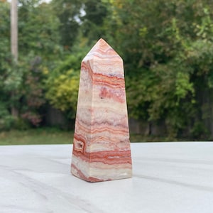 May include: A white, pink, and red banded agate crystal point, also known as a wand or obelisk, is a natural stone used for healing and meditation.