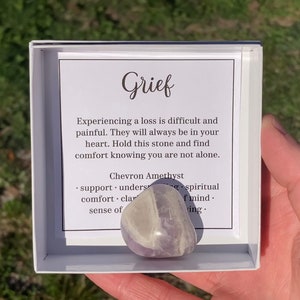 May include: A small white box with a purple chevron amethyst crystal inside. The box has a message that reads "Grief. Experiencing a loss is difficult and painful. They will always be in your heart. Hold this stone and find comfort knowing you are not alone. Chevron Amethyst. support understanding. comfort clarity. sense of peace. spiritual healing. peace of mind. loving."