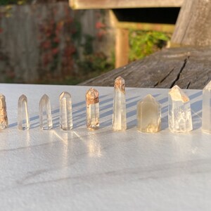 May include: A collection of ten clear quartz crystal points with a variety of sizes and shapes. The crystals are arranged in a row on a white surface.