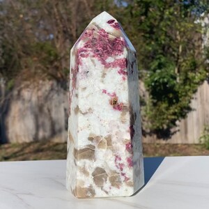 May include: A white and pink crystal point with a pointed top and a flat base. The crystal has a natural, rough texture and is speckled with pink and brown inclusions.