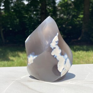 May include: A polished, freeform agate stone with a gray base color and white and blue cloud-like patterns. The stone is displayed on a white surface with a blurred green background of trees and grass.