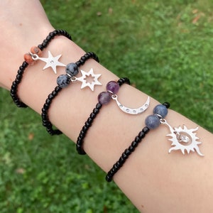 May include: Four black beaded bracelets with silver star, moon, and sun charms. The bracelets feature various gemstone beads, including a light pink, gray, purple, and blue.