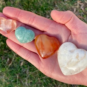 May include: Four heart-shaped polished stones in various colors, including orange, white, green, and pink. The stones are arranged in a row on a hand.