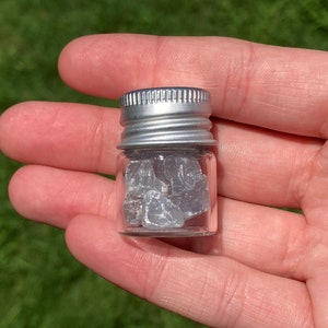 May include: A small glass jar with a silver lid filled with small, gray, iridescent crystals.