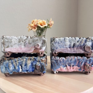 May include: Four ceramic rectangular planters with a crackled glaze finish. The planters are stacked in pairs, with the top two planters in a white and pink glaze and the bottom two planters in a blue and pink glaze. Each planter has a small brown square stamp on the side.