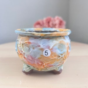 May include: A small, colorful ceramic planter with a textured surface and three legs. The planter has a light blue, yellow, orange, and pink glaze. The number "5" is written on a white circle on the side of the planter.