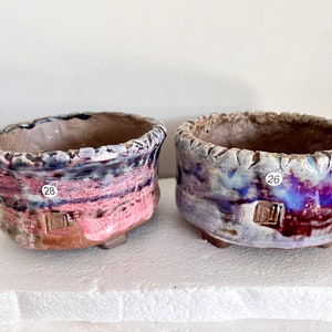 May include: Two small ceramic pots with a textured rim and a glaze that is a mix of pink, purple, and blue. The pots have numbers 26 and 28 written on them.