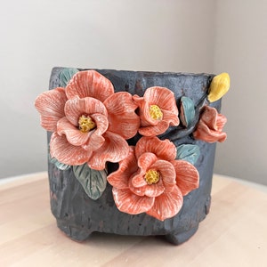 May include: A dark gray ceramic planter with a textured surface, decorated with three dimensional pink flowers and green leaves.