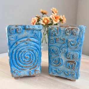 May include: Two blue ceramic planters with different embossed designs. One planter has a spiral pattern and the other has a geometric pattern.