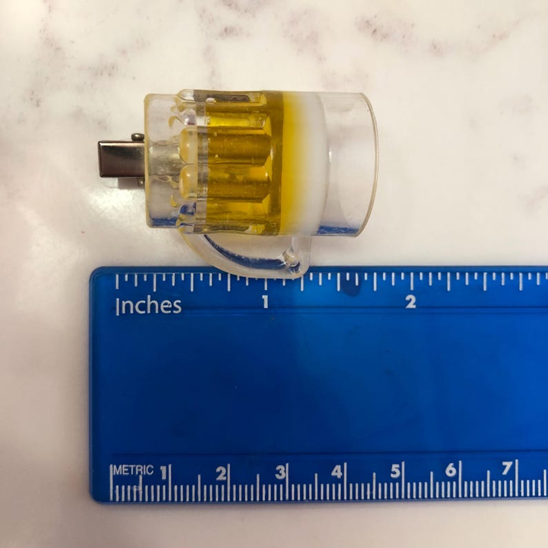 May include: A clear plastic beer mug keychain with a yellow liquid inside. The keychain is about 1 inch long and is measured against a ruler.
