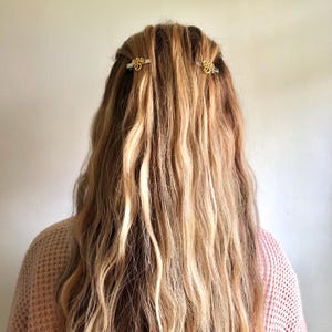 May include: Long, wavy blonde and brown hair styled with two gold-coloured hair clips. The clips have a decorative design and are positioned on either side of the head. The person is wearing a light pink jumper.