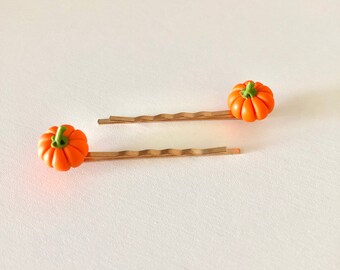 May include: Two gold hair clips with small orange pumpkin charms. The pumpkins have green stems.