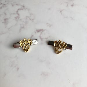 May include: Two gold dragon hair clips with silver rectangular bases. The dragons have detailed wings and bodies. The clips are displayed on a white and grey marble surface.