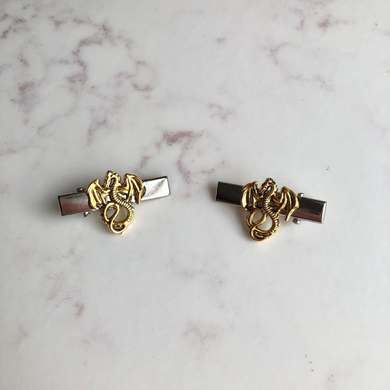 May include: Two gold dragon hair clips with silver rectangular bases. The dragons have detailed wings and bodies. The clips are displayed on a white and grey marble surface.