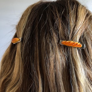 May include: Two silver hair clips with a baguette design. The baguette is a light brown color with a slightly orange hue.