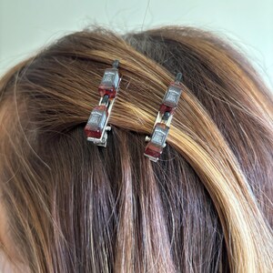 May include: Two silver hair clips, each adorned with three miniature brown and red bottle replicas. The hair accessories are set against a backdrop of brown and blonde hair.