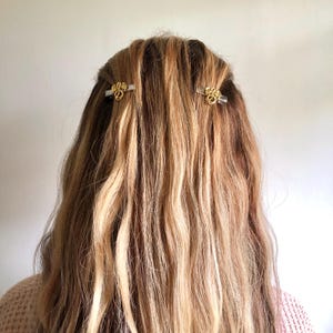 May include: Two gold-coloured hair clips with ornate designs are placed in wavy, light brown and blonde hair. The clips are positioned on either side of the head, adding a touch of elegance to the hairstyle. The background is a plain, off-white colour.