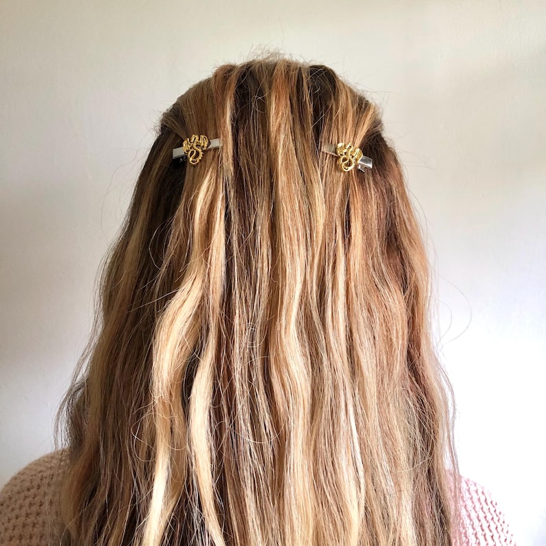 May include: Two gold-coloured hair clips with ornate designs are placed in wavy, light brown and blonde hair. The clips are positioned on either side of the head, adding a touch of elegance to the hairstyle. The background is a plain, off-white colour.
