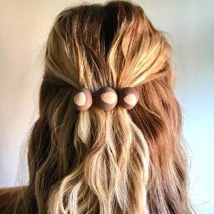 May include: Three brown hair clips with a light pink center are holding back wavy blonde and brown hair. The hair clips are round and evenly spaced. The hair is styled in a half-up, half-down style.