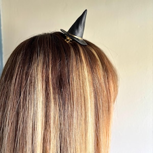 May include: A black witch hat hair clip with a gold band and clasp, perched atop long, highlighted hair. The miniature hat is a whimsical accessory, perfect for Halloween or themed events. The hair has blonde and brown highlights.