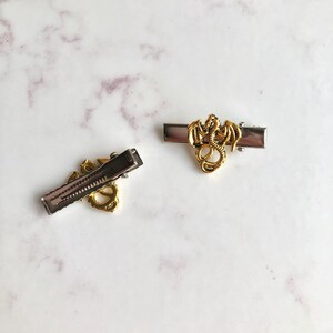 May include: Two metal hair slides featuring a gold dragon design. The slides have a silver base and a gold dragon figure. The dragon is detailed with wings and a curled tail. The slides are on a white and grey marble surface.