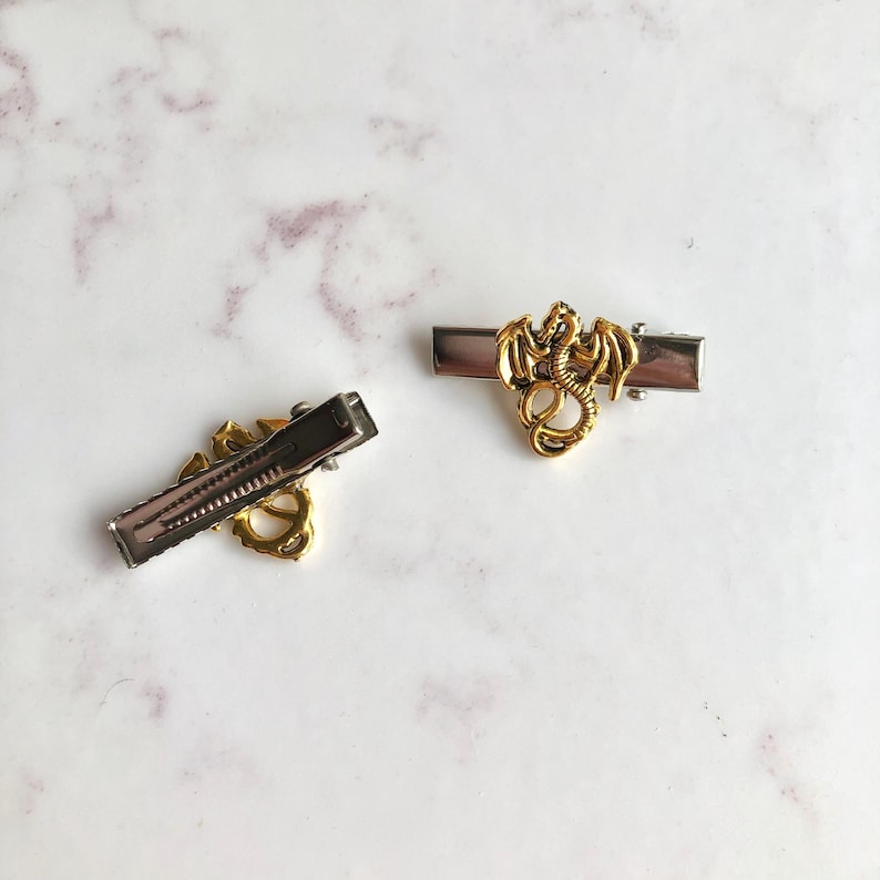May include: Two metal hair slides featuring a gold dragon design. The slides have a silver base and a gold dragon figure. The dragon is detailed with wings and a curled tail. The slides are on a white and grey marble surface.