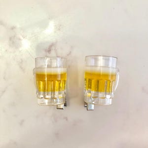 May include: Two clear glass beer mugs with a yellow liquid inside. The mugs have a handle and are attached to a metal clip.