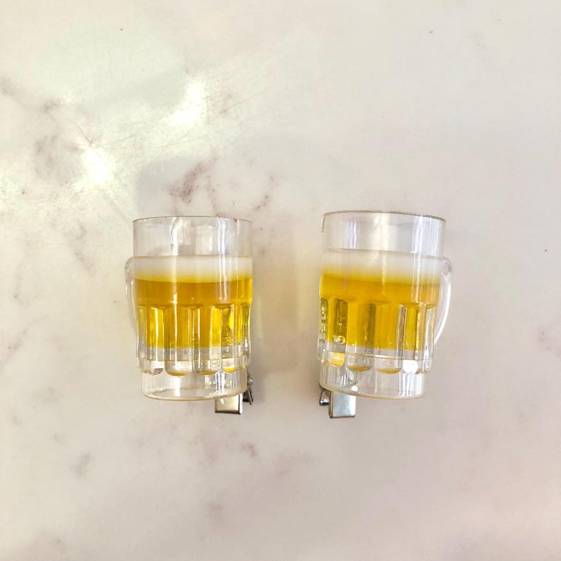 May include: Two clear glass beer mugs with a yellow liquid inside. The mugs have a handle and are attached to a metal clip.
