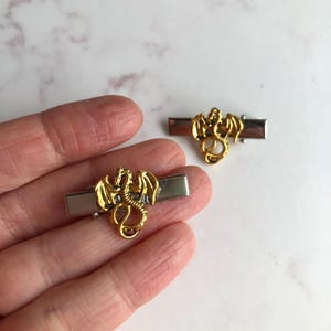 May include: Two gold-coloured dragon tie clips. The dragon has detailed wings and a curled tail. The tie clips have a silver-coloured bar. The dragon is centered on the bar.