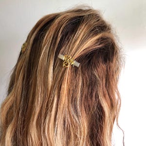 May include: Close-up of hair with gold-coloured hair clips. The clips have a rectangular silver base with an ornate gold design. The hair is wavy and has shades of brown and blonde.