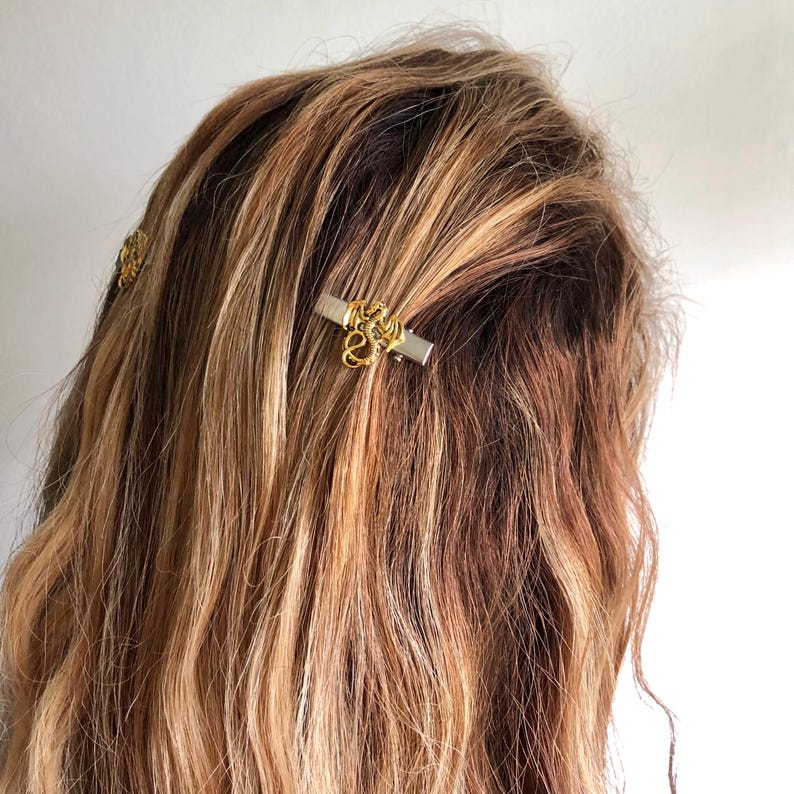 May include: Close-up of hair with gold-coloured hair clips. The clips have a rectangular silver base with an ornate gold design. The hair is wavy and has shades of brown and blonde.