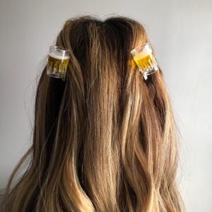 May include: Two clear plastic hair clips shaped like shot glasses filled with a yellow liquid, resembling beer. The clips are in a person's blonde hair.