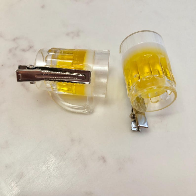 May include: Two clear plastic beer mug hair clips with a yellow liquid inside. The clips are silver and have a spring mechanism.