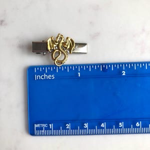 May include: A gold dragon tie clip with a silver clip. The dragon is approximately 2.5 cm long. The clip is on a white surface with a blue ruler that has both inches and metric measurements.